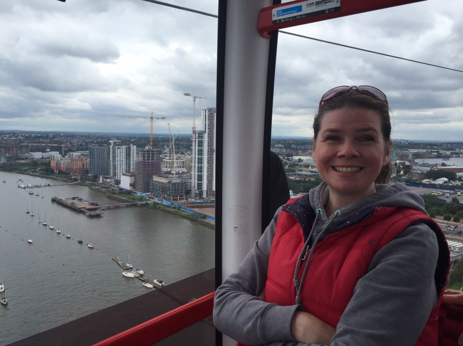 I'm in a pod on a cable car high above the River Thames. Boats, houses and a blocks of flats are below me. I'm wearing a red gilet and grey hoodie, my arms are folded and I am grinning at the camera.